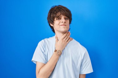 Mavi arka planda dikilen İspanyol genç adam ağrılı boynuna dokunuyor. Grip, mantar ve enfeksiyon yüzünden boğazı ağrıyor. 