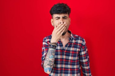 Kırmızı arka planda dikilen sakallı genç İspanyol adam ağzını eliyle kapatmaktan sıkılmış. Huzursuz ve uykulu.. 