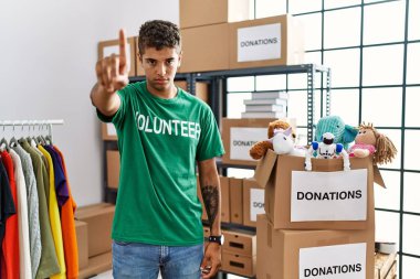 Young handsome hispanic man wearing volunteer t shirt at donations stand pointing with finger up and angry expression, showing no gesture 