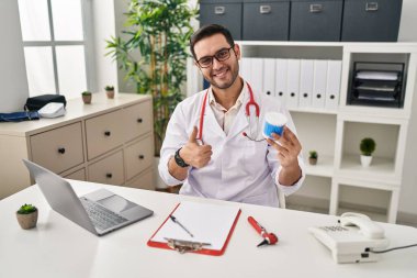 Sakallı, pamuklu sakallı, mutlu ve pozitif gülümseyen genç İspanyol doktor başparmağıyla mükemmel ve onay işareti yapıyor. 