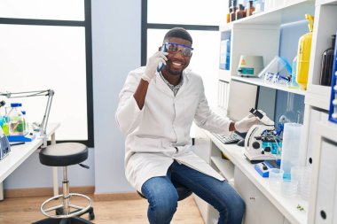 African american man working at scientist laboratory speaking on the phone looking positive and happy standing and smiling with a confident smile showing teeth 