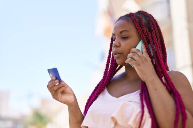 African american woman talking on the smartphone and using credit card at street