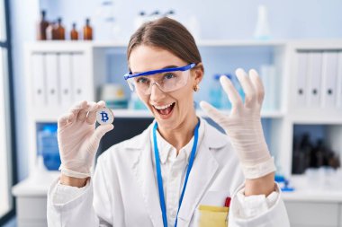 Young caucasian woman working at scientist laboratory holding bitcoin annoyed and frustrated shouting with anger, yelling crazy with anger and hand raised 