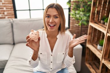 Genç sarışın kadın kumbarayı tutuyor başarıyı kutluyor mutlu bir gülümsemeyle ve kazanan yüz ifadesiyle elini kaldırarak. 