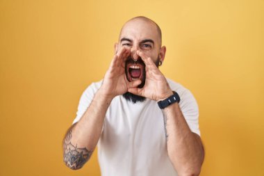Young hispanic man with beard and tattoos standing over yellow background shouting angry out loud with hands over mouth 