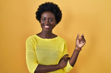 African young woman standing over yellow studio with a big smile on face, pointing with hand finger to the side looking at the camera. 