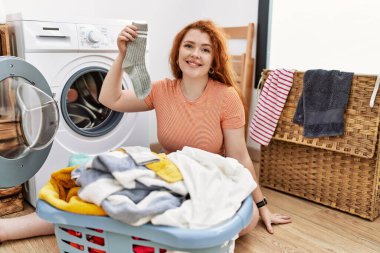 Young redhead woman putting dirty laundry into washing machine with a happy and cool smile on face. lucky person. 