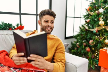 Genç Arap adam kitap okuyor. Evdeki Noel ağacının yanındaki kanepede yatıyor..
