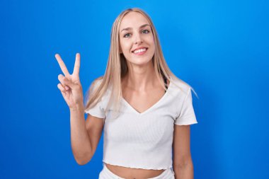 Young caucasian woman standing over blue background showing and pointing up with fingers number two while smiling confident and happy. 