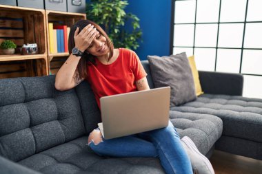 Young hispanic woman using laptop at home stressed and frustrated with hand on head, surprised and angry face 