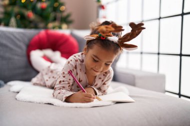 Adorable hispanic girl drawing on notebook lying on sofa by christmas tree at home