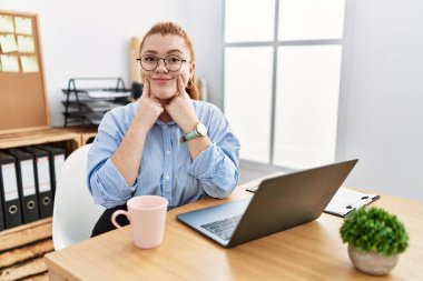 Young redhead woman working at the office using computer laptop smiling with open mouth, fingers pointing and forcing cheerful smile 