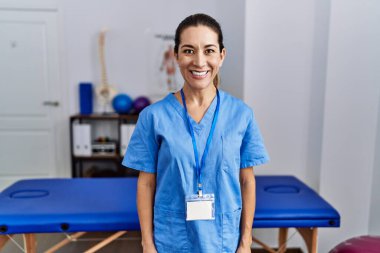 Young hispanic woman wearing physiotherapist uniform standing at clinic with a happy and cool smile on face. lucky person. 