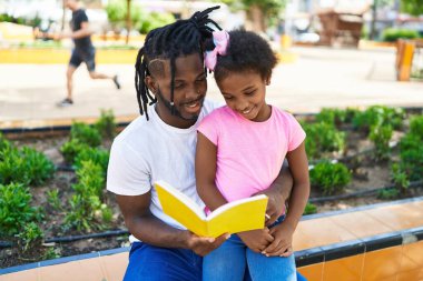 Father and daughter reading book sitting together on bench at park