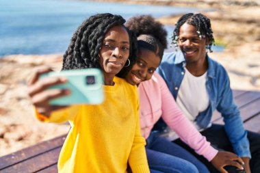 African american friends making selfie by the smartphone sitting on bench at seaside