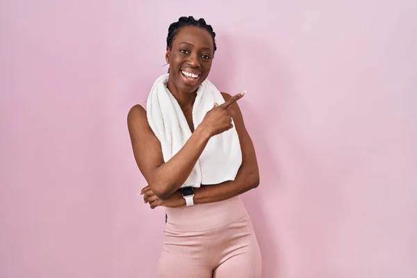 Beautiful black woman wearing sportswear and towel over pink background with a big smile on face, pointing with hand and finger to the side looking at the camera.