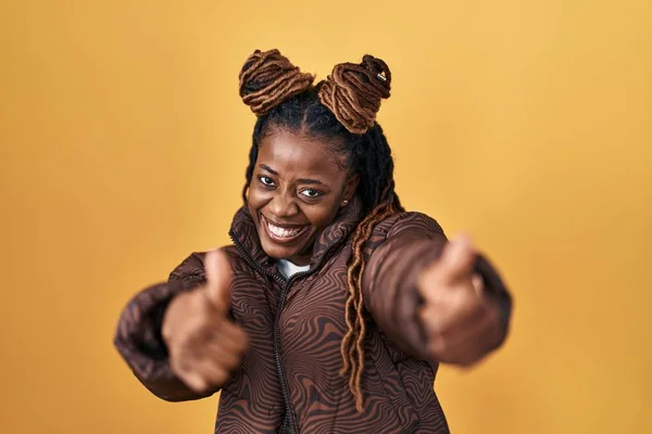 African woman with braided hair standing over yellow background approving doing positive gesture with hand, thumbs up smiling and happy for success. winner gesture.