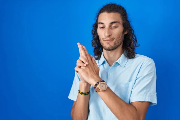 Young hispanic man standing over blue background holding symbolic gun with hand gesture, playing killing shooting weapons, angry face