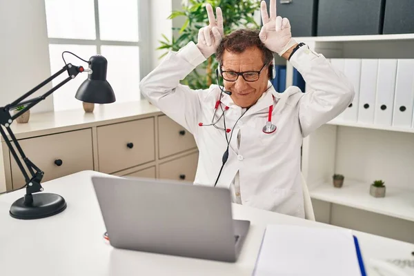 Senior doctor man working on online appointment posing funny and crazy with fingers on head as bunny ears, smiling cheerful