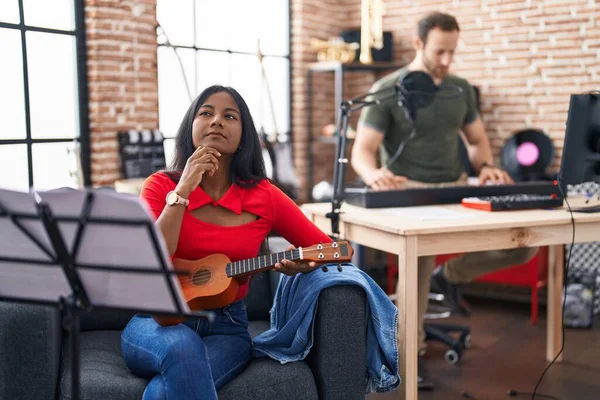 Young indian woman playing ukulele at music studio serious face thinking about question with hand on chin, thoughtful about confusing idea