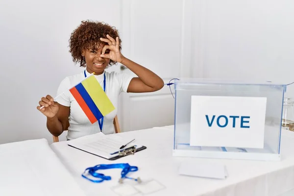 Young african american woman at political campaign election holding colombia flag smiling happy doing ok sign with hand on eye looking through fingers