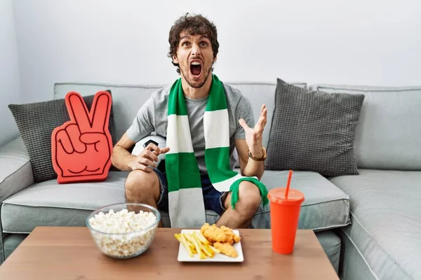 Young hispanic man football hooligan cheering game at home crazy and mad shouting and yelling with aggressive expression and arms raised. frustration concept.
