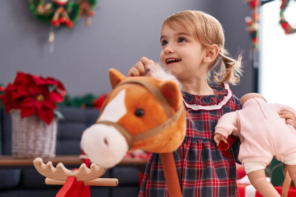 愛らしいですCaucasian女の子保持赤ちゃん人形と馬おもちゃスタンディングによってクリスマス装飾で家 — ストック写真