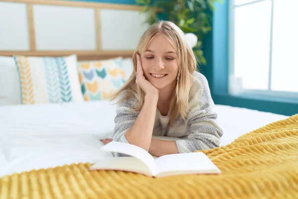 Young blonde woman reading book lying on bed at bedroom