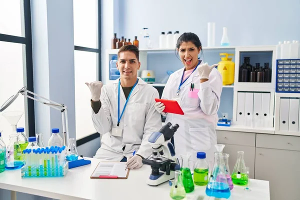 Young doctors working at scientist laboratory pointing thumb up to the side smiling happy with open mouth