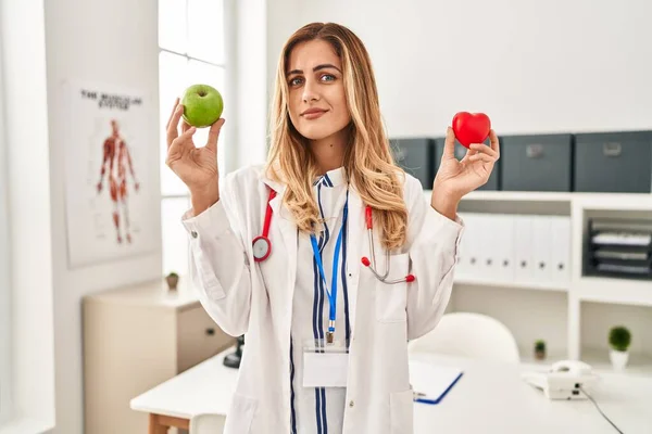 Young Blonde Doctor Woman Holding Heart Green Apple Clueless Confused — Stok fotoğraf