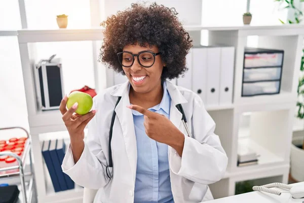 Black doctor woman with curly hair holding green apple smiling happy pointing with hand and finger