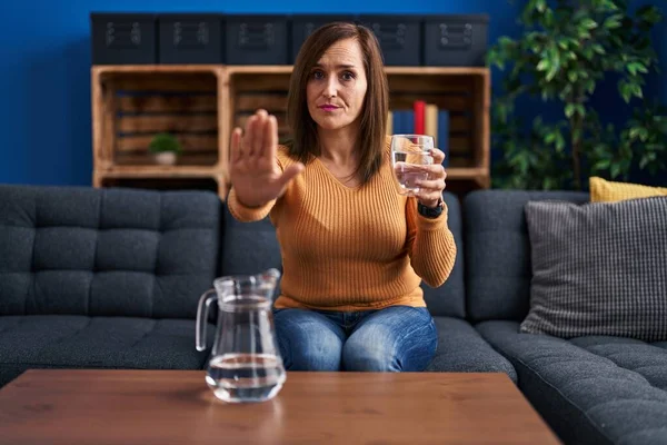 Middle age woman drinking glass of water with open hand doing stop sign with serious and confident expression, defense gesture