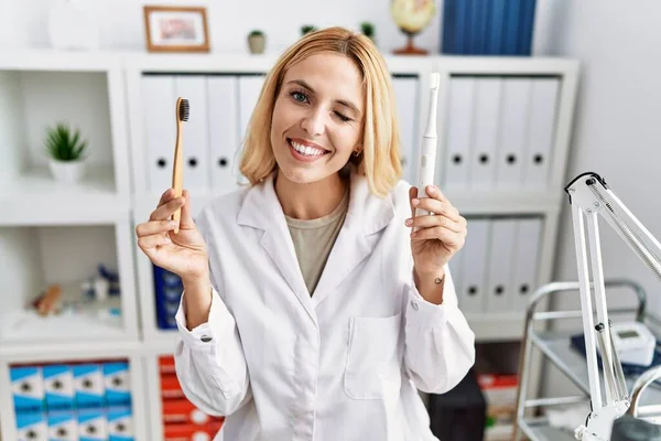Beautiful Blonde Dentist Woman Holding Electric Toothbrush Normal Toothbrush Winking — Stockfoto