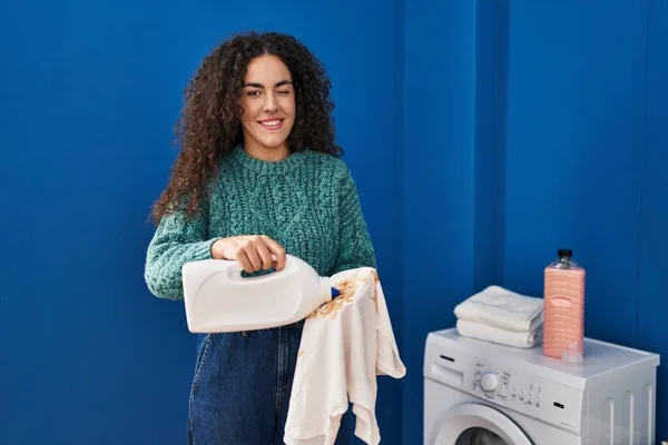 Young Hispanic Woman Holding Dirty Laundry Detergent Bottle Winking Looking — Stockfoto