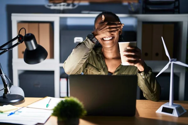 African woman working using computer laptop at night smiling and laughing with hand on face covering eyes for surprise. blind concept.