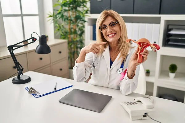 Middle age blonde gynecologist woman holding anatomical model of female genital organ pointing finger to one self smiling happy and proud