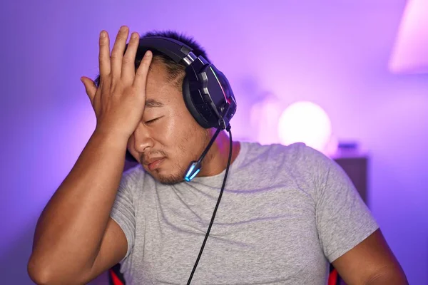 Chinese young man playing video games wearing headphones surprised with hand on head for mistake, remember error. forgot, bad memory concept.