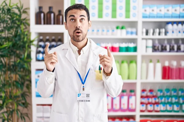 Красивый Латиноамериканец Работающий Аптеке Поражен Удивлен Глядя Вверх Указывая Пальцами — стоковое фото