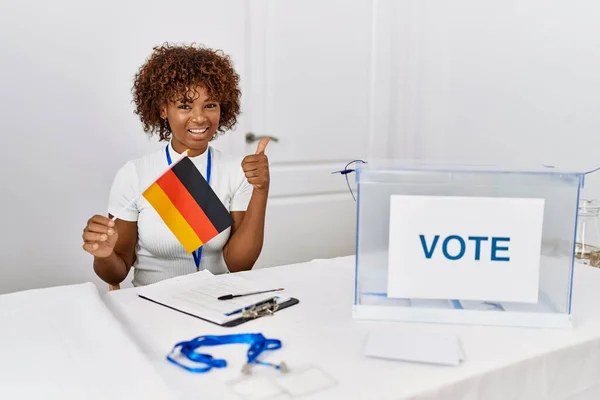 Young african american woman at political campaign election holding germany flag smiling happy and positive, thumb up doing excellent and approval sign