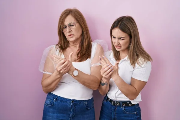 Hispanic mother and daughter wearing casual white t shirt over pink background suffering pain on hands and fingers, arthritis inflammation