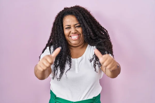 ピンクの背景の上に立つプラスサイズのヒスパニック系女性は 手で積極的なジェスチャーを行うことを承認します 親指アップ笑顔と成功のために幸せ 勝者のジェスチャー — ストック写真