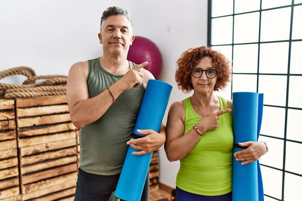 Middle age couple holding yoga mat pointing with hand finger to the side showing advertisement, serious and calm face