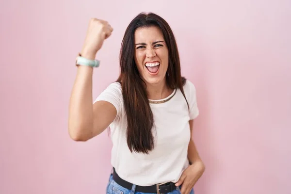 Young brunette woman standing over pink background angry and mad raising fist frustrated and furious while shouting with anger. rage and aggressive concept.