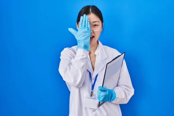 Chinese young woman working at scientist laboratory covering one eye with hand, confident smile on face and surprise emotion.