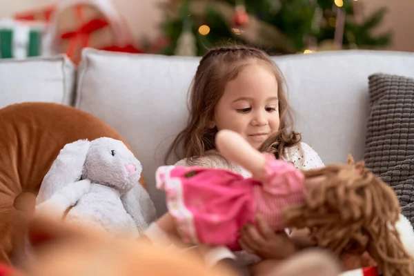 Adorable hispanic girl holding doll sitting on sofa by christmas tree at home