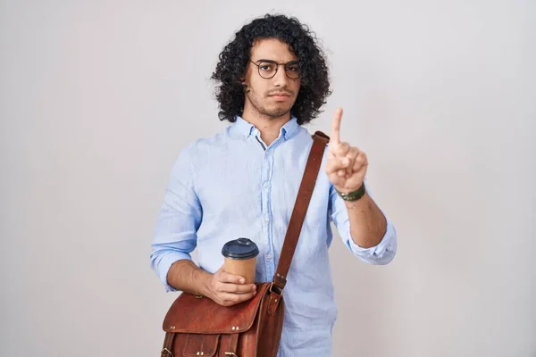 Hispanic man with curly hair drinking a cup of take away coffee pointing with finger up and angry expression, showing no gesture