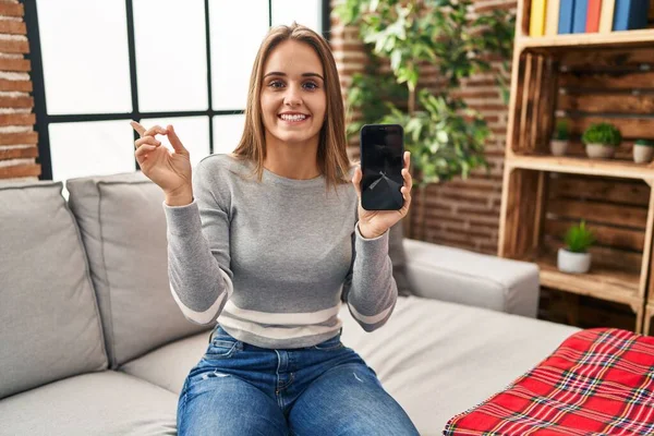 Young woman holding broken smartphone showing cracked screen smiling happy pointing with hand and finger to the side
