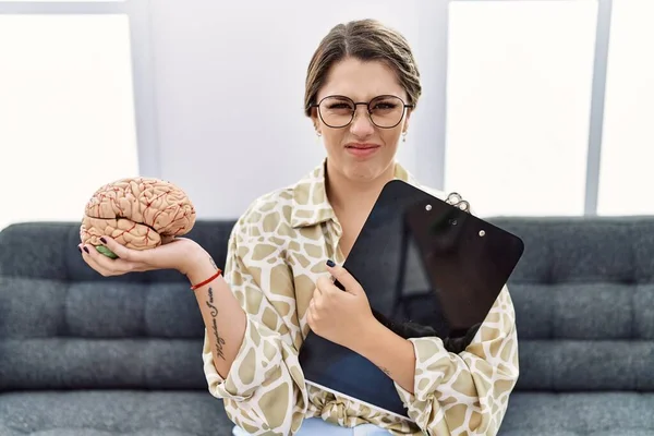 Young hispanic woman holding brain working at psychology clinic clueless and confused expression. doubt concept.