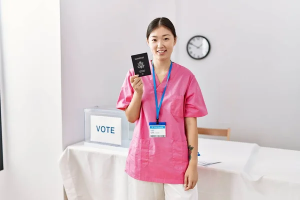 Young asian nurse woman at political campaign election holding usa passport looking positive and happy standing and smiling with a confident smile showing teeth