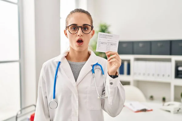 Młody Lekarz Kobieta Gospodarstwa Covid Certyfikat Przestraszony Zdumiony Otwartymi Ustami — Zdjęcie stockowe